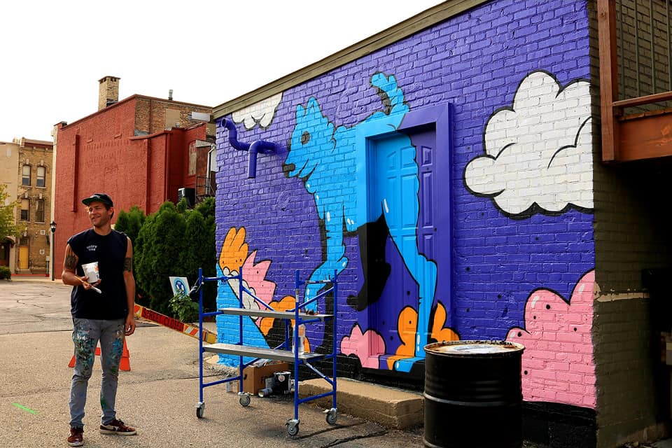 An artist poses in front of a mural he painted during the Downtown Janesville Art Infusion