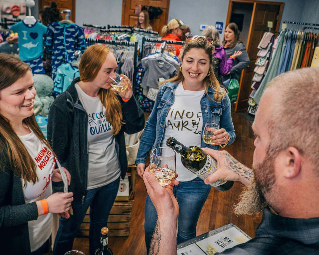 A group of women sip wine inside a downtown store while a man pours a glass of wine