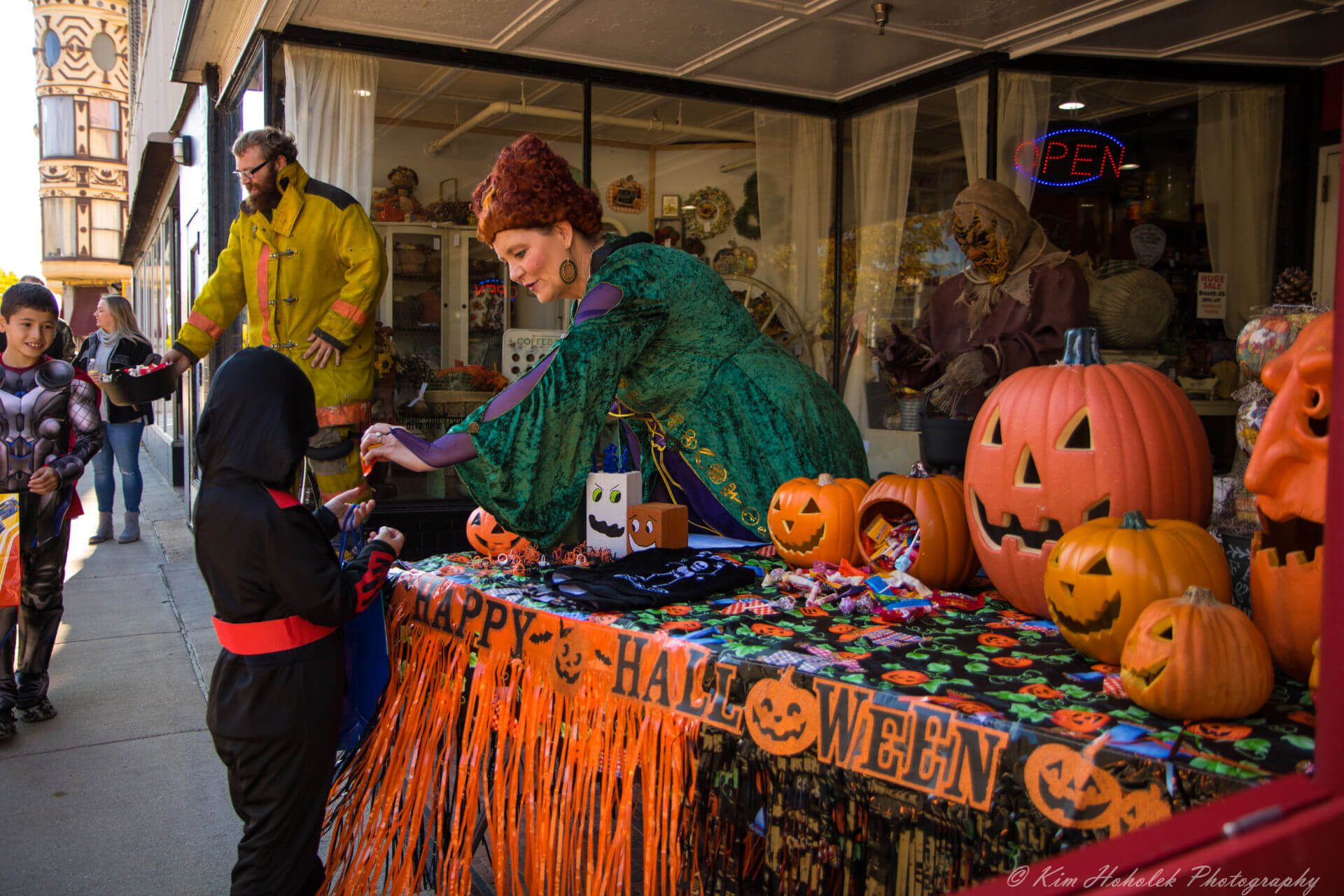 A costumed woman, standing behind a table filled with jack-o-lanterns, hands a piece of candy to a young trick-or-treater in downtown Janesville.
